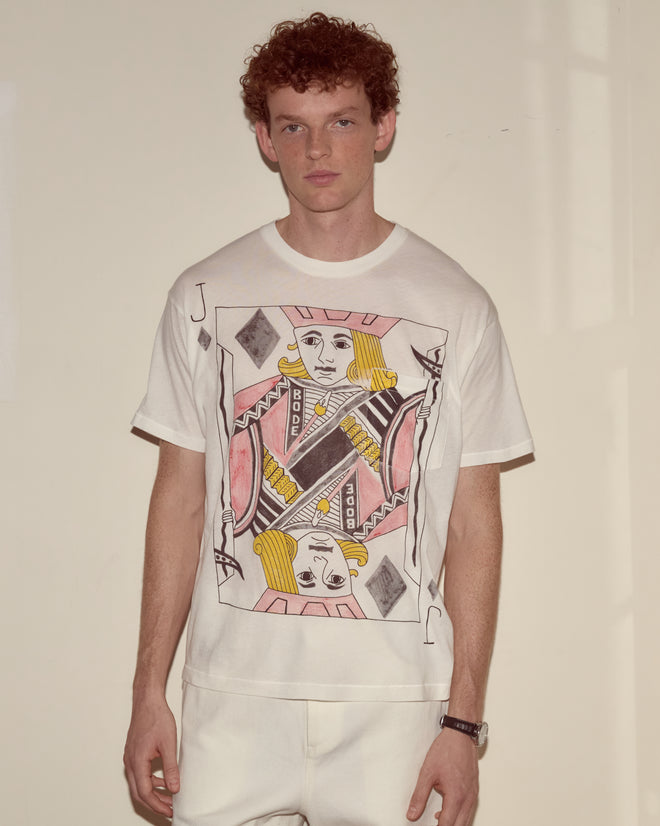 A man in white stands against a beige wall, wearing the Jack Of Diamonds Pocket Tee—100% cotton and inspired by Alice Through the Looking Glass.