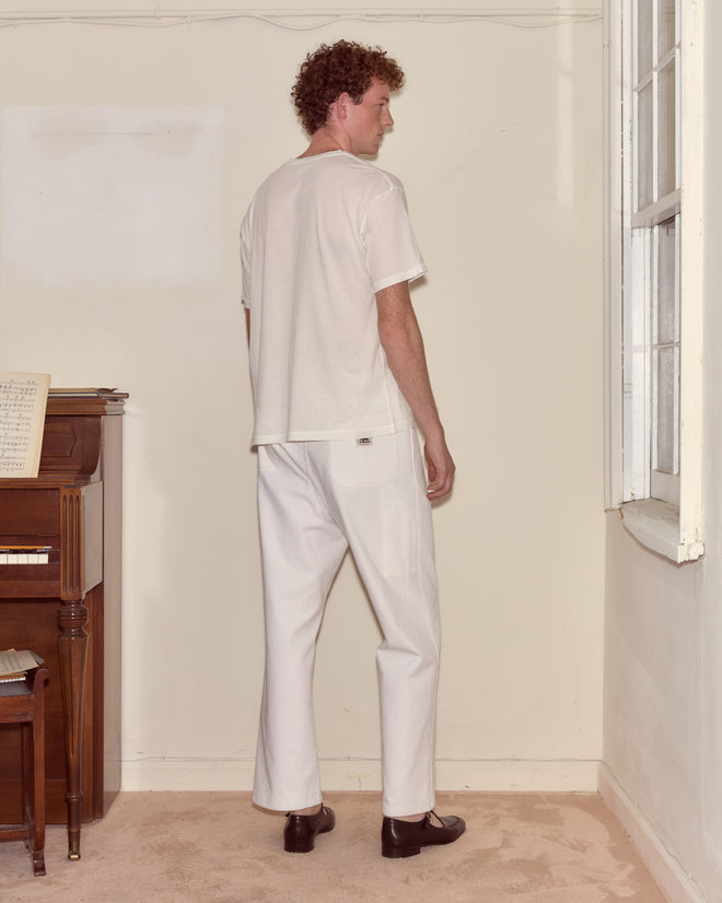 A man wearing a Jack Of Diamonds Pocket Tee and white pants stands indoors near a piano, facing away from the camera, evoking an Alice Through the Looking Glass scene.