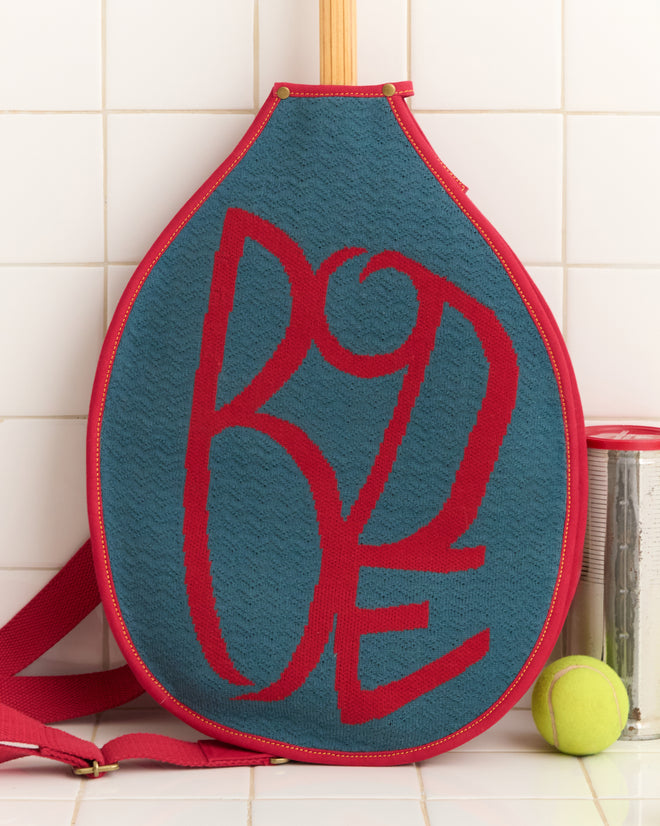 The Jacquard Racquet Sleeve, featuring bold red lettering and an adjustable strap, is displayed alongside a tennis ball and can against a tiled wall.