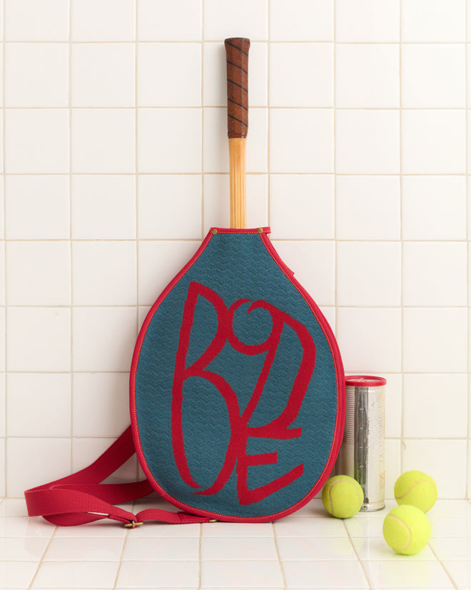 The Jacquard Racquet Sleeve, featuring red lettering and an adjustable strap, rests on a tiled surface next to a can of tennis balls and three loose balls.