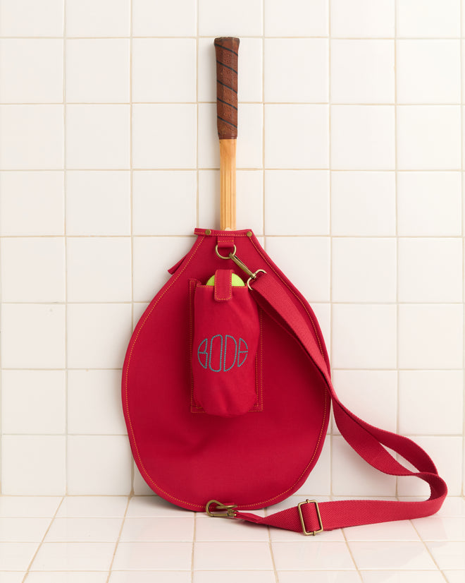 The Jacquard Racquet Sleeve, featuring a red vintage design with an adjustable strap and a small essentials pouch, rests against a white tiled wall.