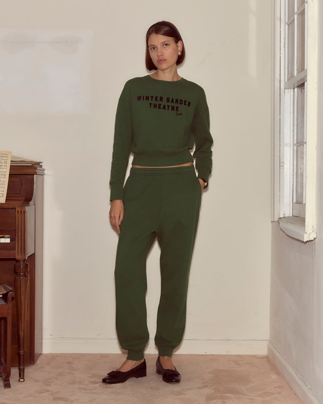 Woman in green "Winter Garden Theatre" sweatshirt made from midweight fleece and matching Joggers - Dark Green stands indoors near a piano and window, the look finished with a signature Bode woven label.