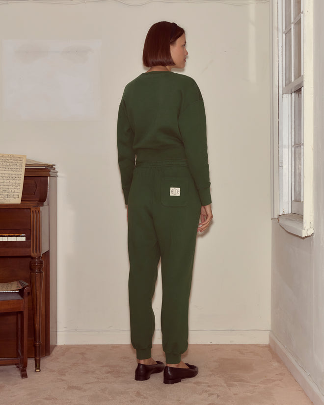 A woman in dark green joggers stands indoors facing a wall, positioned beside a piano and a window.