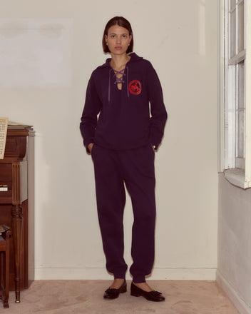 Woman in dark purple joggers stands indoors near a piano and window, hands in pockets, facing forward.