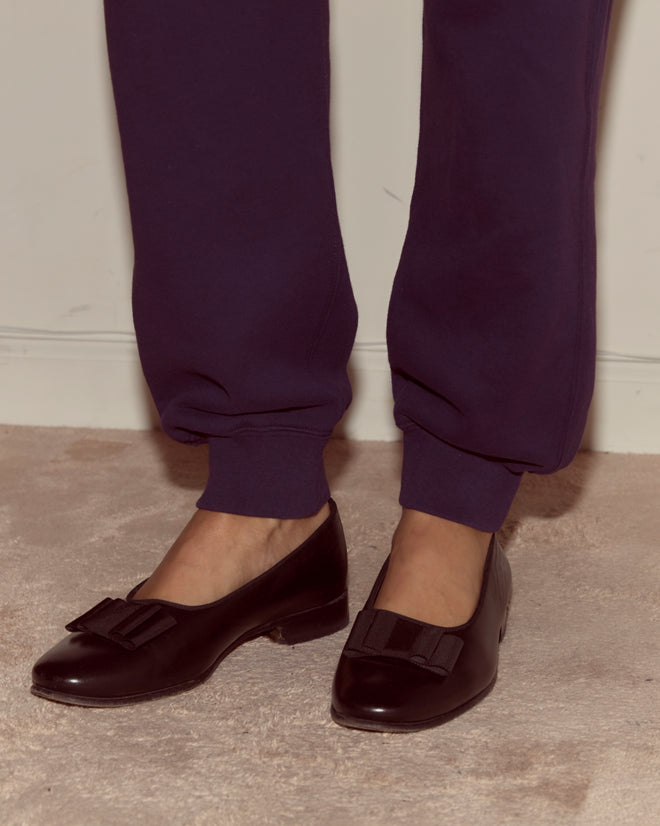 Person wearing dark loafers with bows and Dark Purple Joggers standing on a beige carpet near a white wall.