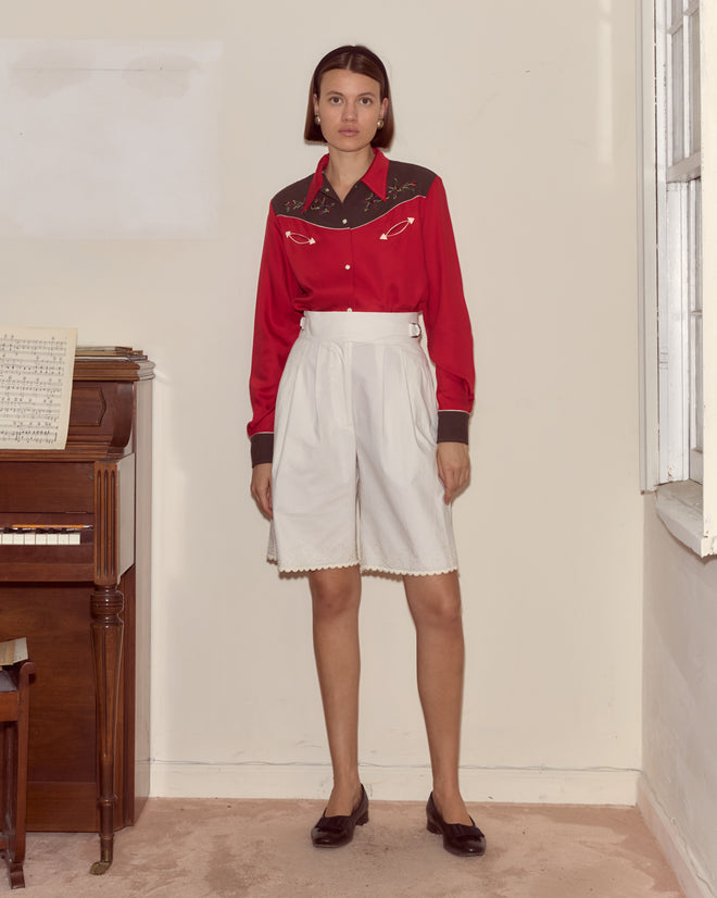 A woman wears the Jumper Western Shirt in red with white shorts, standing indoors by a piano and window, facing the camera.