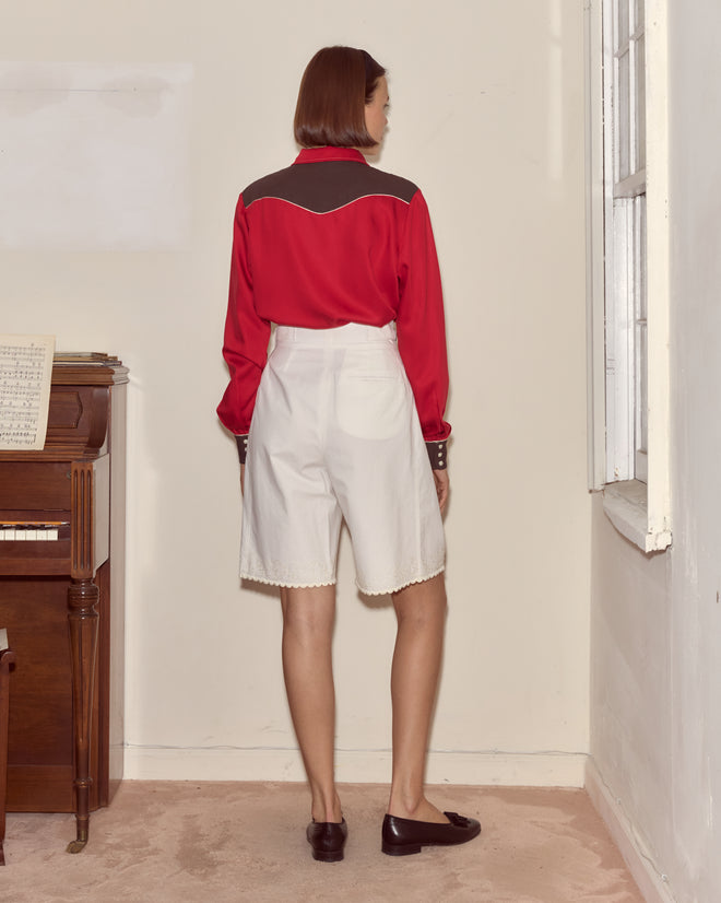 A woman in a Jumper Western Shirt and white shorts stands facing a wall beside a piano in a softly lit room.