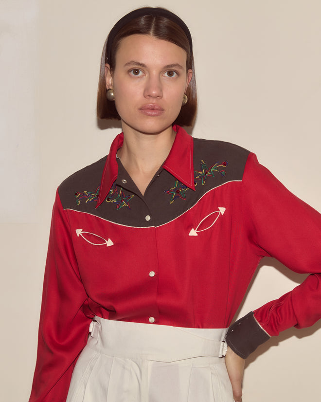 A woman wearing the Jumper Western Shirt, featuring vintage-inspired red and brown floral embroidery, pairs it with white pants and stands against a plain beige background.