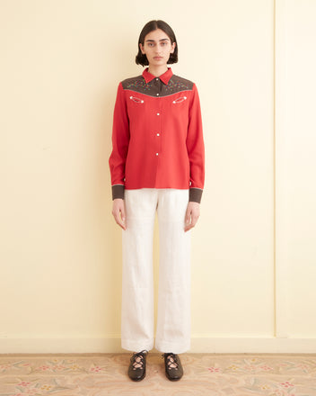 Wearing the Jumper Western Shirt, with its red and brown embroidered star and arrow designs, a person is seen against a light background. The addition of floral motifs enhances the shirt's charm.