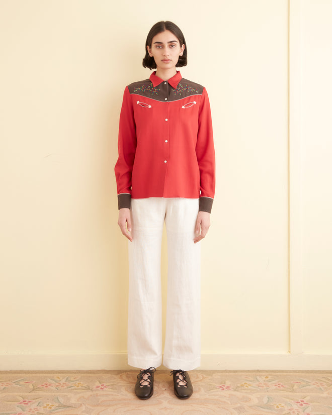 Wearing the Jumper Western Shirt, with its red and brown embroidered star and arrow designs, a person is seen against a light background. The addition of floral motifs enhances the shirt's charm.