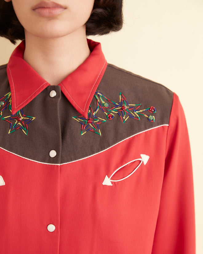 Wearing the Jumper Western Shirt, with its red and brown embroidered star and arrow designs, a person is seen against a light background. The addition of floral motifs enhances the shirt's charm.