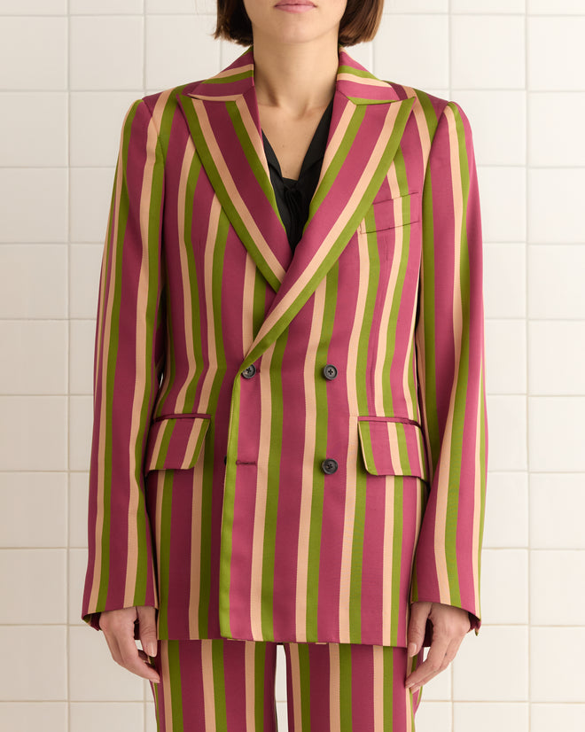 A person stands against a tiled wall, wearing the vibrant Juniper Stripe Double-Breasted Suit Jacket made from Kutnu fabric with pink, green, and yellow stripes.