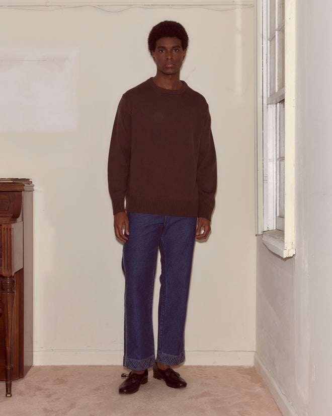 A man wearing the Juniper Sweater in Brown/Black and blue jeans stands indoors by a window and a piano.