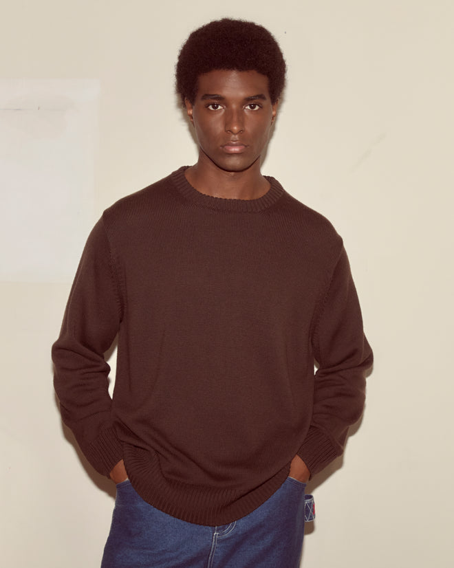 A man in a Juniper Sweater - Brown/Black and blue jeans stands against a plain beige wall with his hands in his pockets.