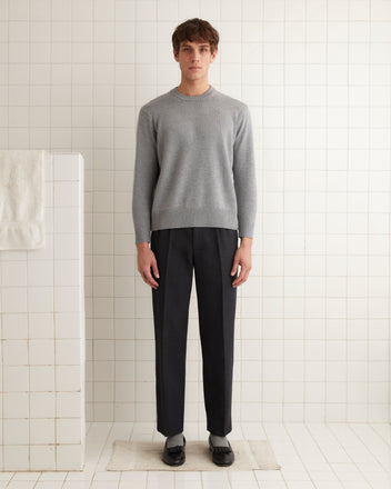 A man in a vintage Juniper Sweater - Grey and dark pants stands on a mat against a tiled wall.