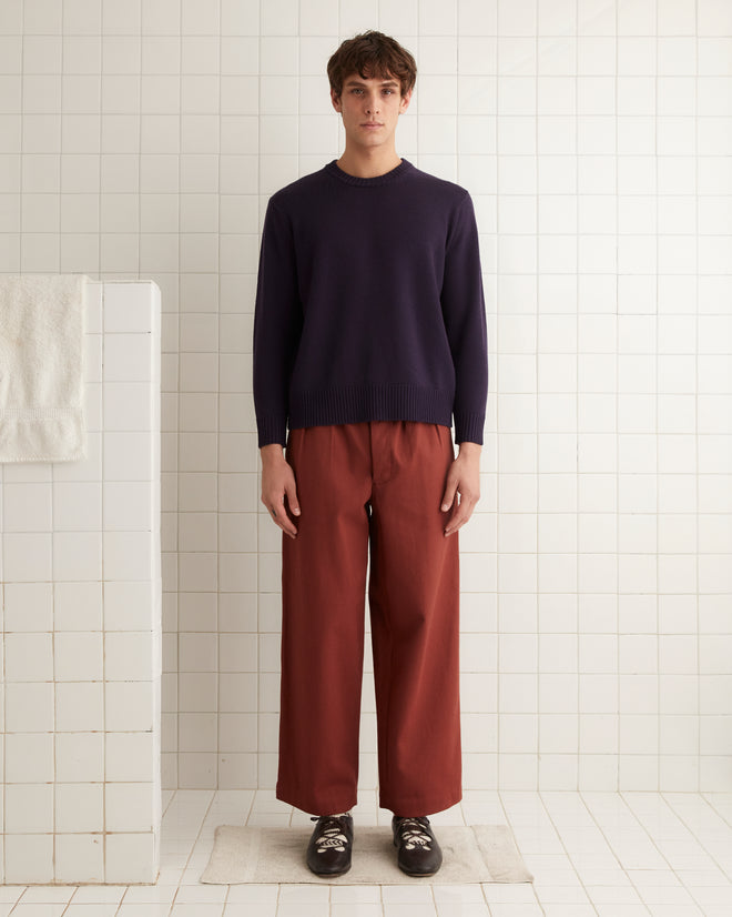 A person in a dark purple Juniper Sweater with ribbed detailing and rust-colored pants stands on a tiled floor with a towel hanging nearby.