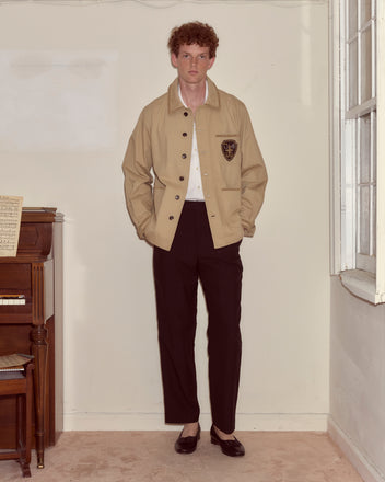 A person wearing the Khaki Bullion Jacket and black pants stands indoors beside a piano near a window.