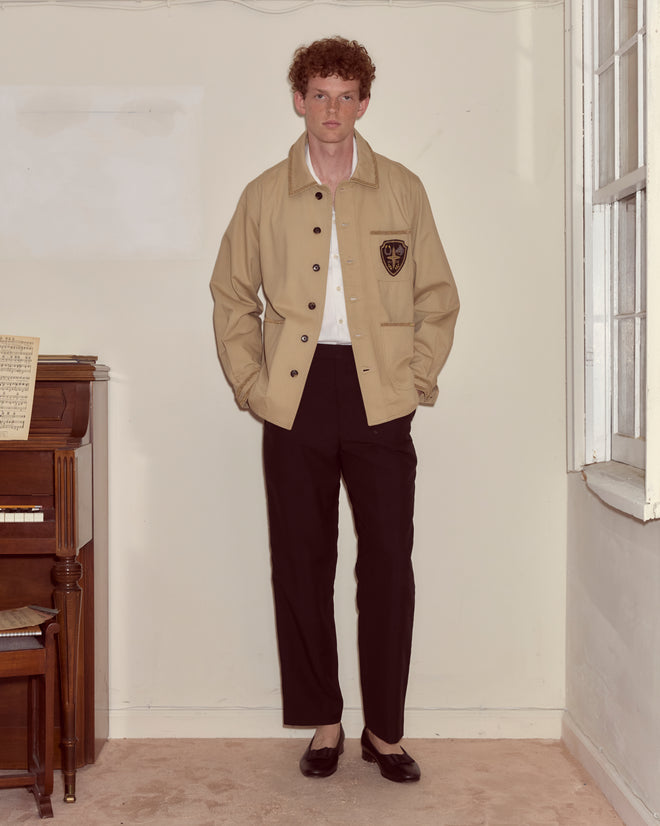 A person wearing the Khaki Bullion Jacket and black pants stands indoors beside a piano near a window.