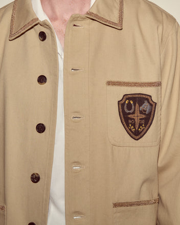 Person wearing the Khaki Bullion Jacket, featuring dark buttons and an embroidered crest patch on the chest pocket.