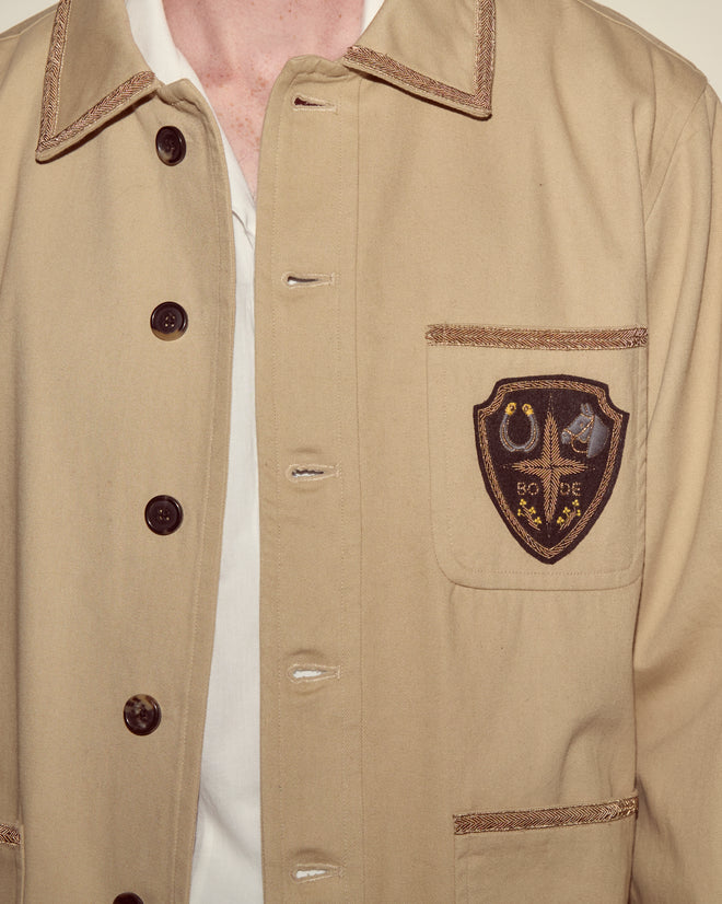 Person wearing the Khaki Bullion Jacket, featuring dark buttons and an embroidered crest patch on the chest pocket.