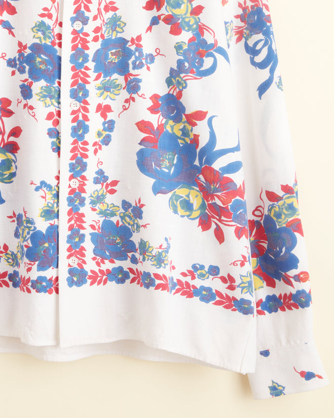 The Protea Long Sleeve Shirt - S/M, featuring unique white fabric adorned with vibrant blue, red, and green florals, hangs on a hook against a light-colored wall.