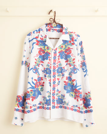 The Protea Long Sleeve Shirt - S/M, featuring unique white fabric adorned with vibrant blue, red, and green florals, hangs on a hook against a light-colored wall.