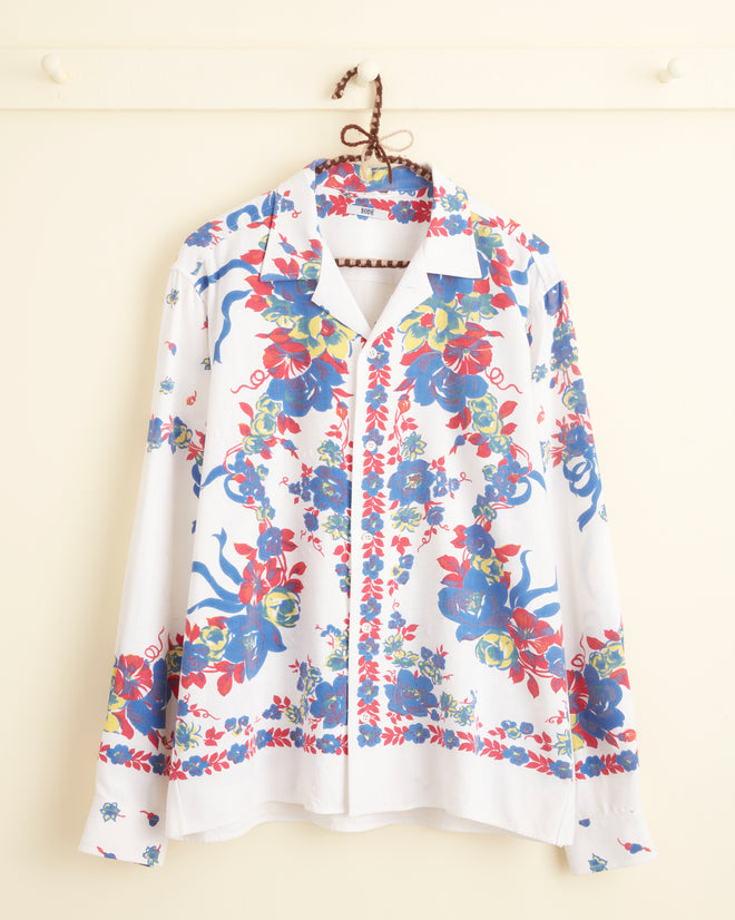 The Protea Long Sleeve Shirt - S/M, featuring unique white fabric adorned with vibrant blue, red, and green florals, hangs on a hook against a light-colored wall.