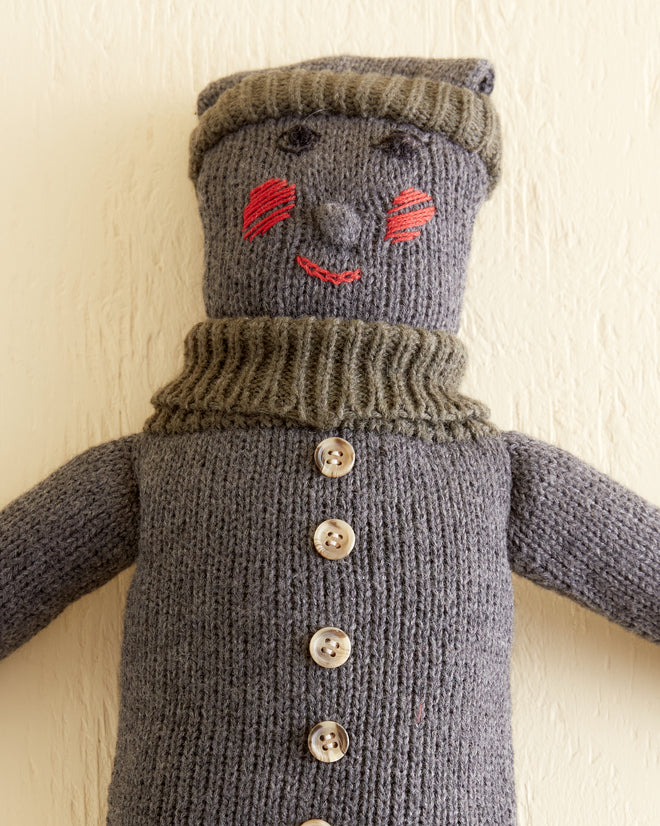 The Knit Doll - Grey Multi is a handmade knitted doll, reminiscent of a 1950s design, adorned in a grey sweater and hat, with red-stitched shoes and rosy cheeks. This timeless piece lies against a beige background and is crafted from 100% wool.