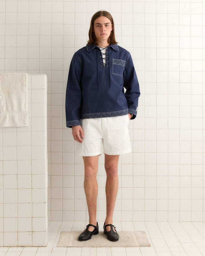 A person in an Embroidered Knolly Brook Pullover - Indigo and white shorts stands on a mat in a tiled room, clutching a towel.