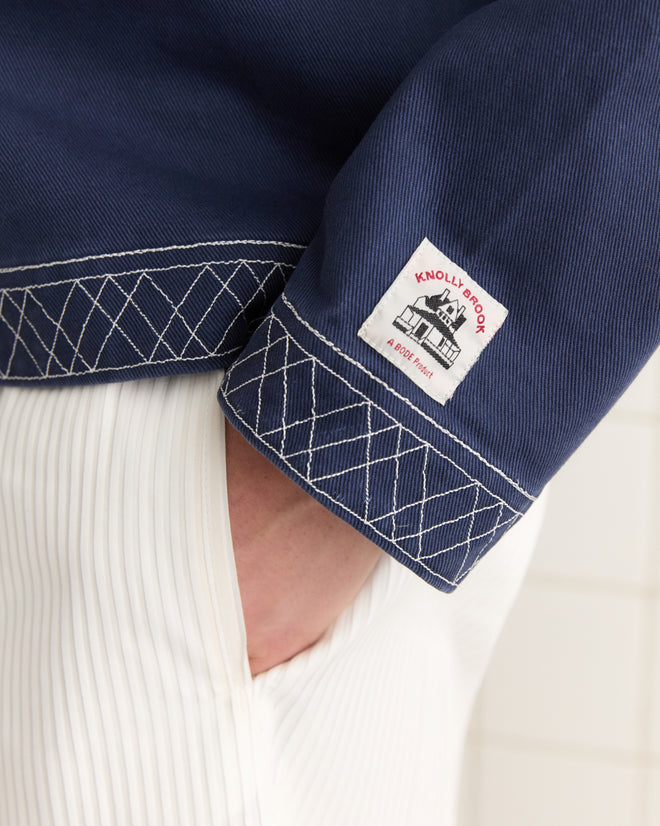 Close-up of a person wearing the Embroidered Knolly Brook Pullover in Indigo, featuring white stitching, a lace-up neckline, and a label that reads "Knolly Brook, A 007 Hotel.