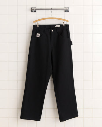 The Twill Knolly Brook Trousers in black, inspired by 1950s–1960s style, hang on a wooden hanger against a white tiled wall.