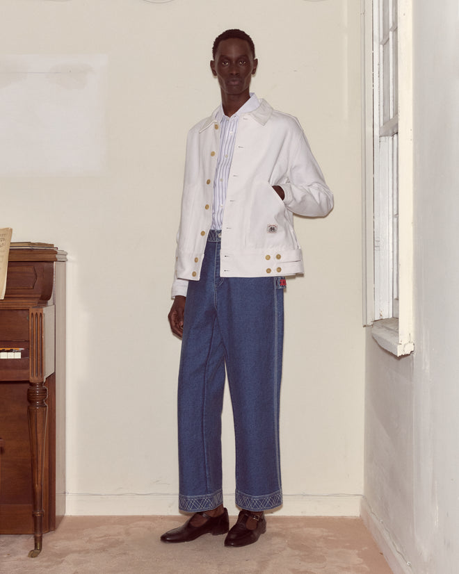 A man stands in a room wearing the Knolly Brook Side Tab Jacket in White, channeling the rugged style of classic workwear labels.