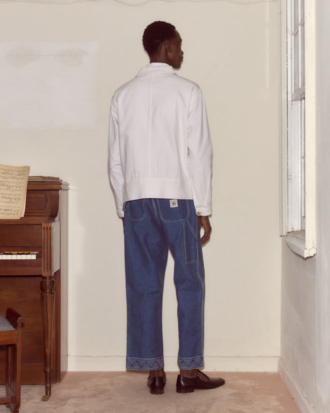 Wearing the Knolly Brook Side Tab Jacket in White and blue jeans, a person stands facing a wall beside a piano in a light-colored room, channeling classic workwear style.