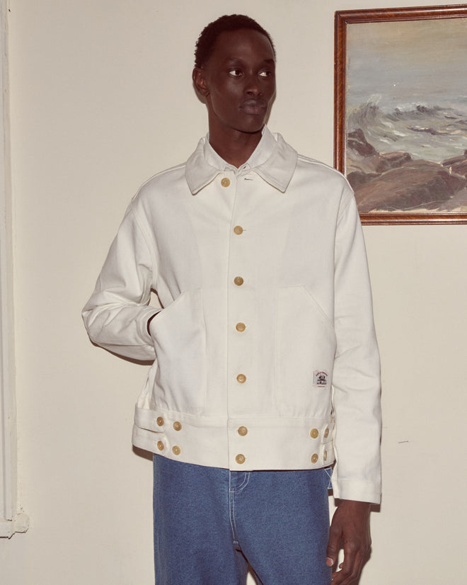 Wearing the Knolly Brook Side Tab Jacket in white with blue jeans, a person stands indoors near a framed painting of the sea.