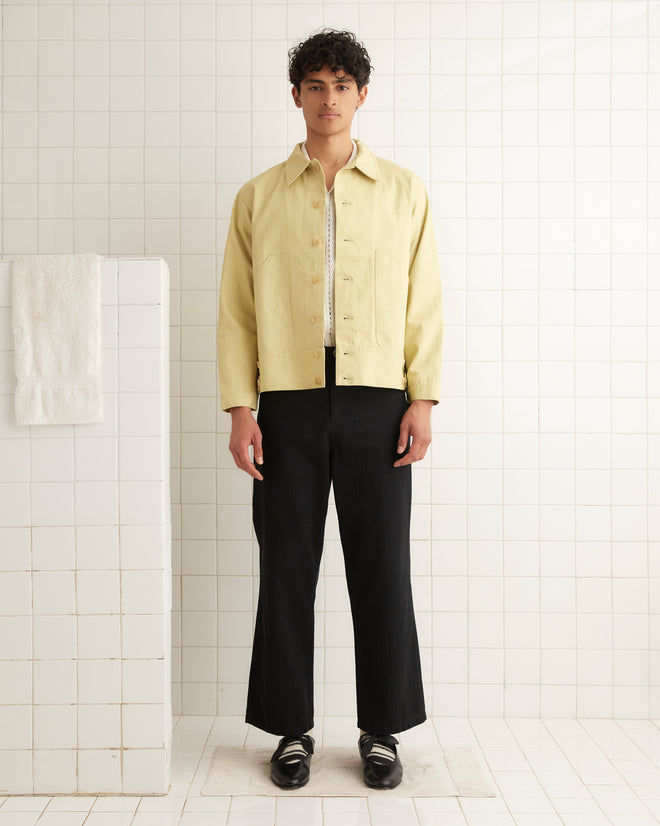 A person dressed in the Knolly Brook Side Tab Jacket - Green and black pants stands in a tiled room next to a white towel, exuding a casual elegance reminiscent of classic workwear style.