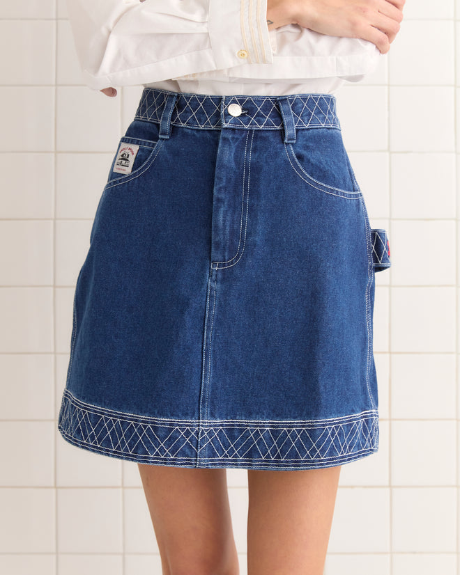 A person wearing an Embroidered Denim Skirt with white stitching, adorned with subtle workwear labels, stands against a tiled wall.