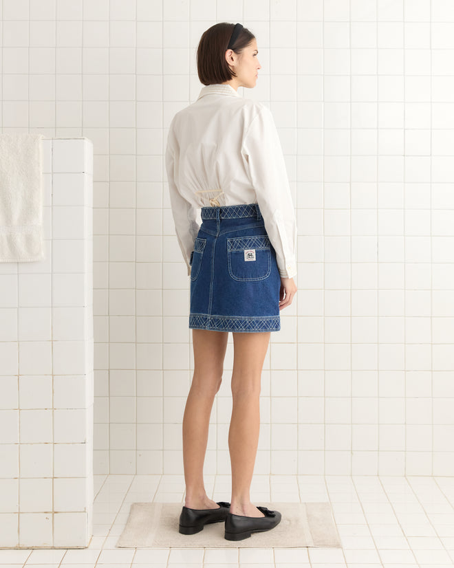 In a tiled room, someone wearing a white shirt and an Embroidered Denim Skirt stands facing away with a towel on the wall.