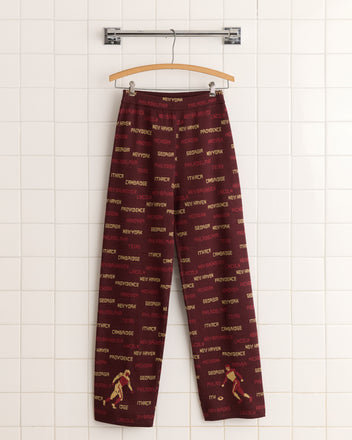 Gridiron Joggers, in bold red with city names artfully printed, feature a wide-leg silhouette. Made from 100% cotton, they hang stylishly on a tile wall.