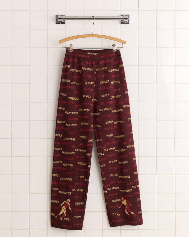 Gridiron Joggers, in bold red with city names artfully printed, feature a wide-leg silhouette. Made from 100% cotton, they hang stylishly on a tile wall.