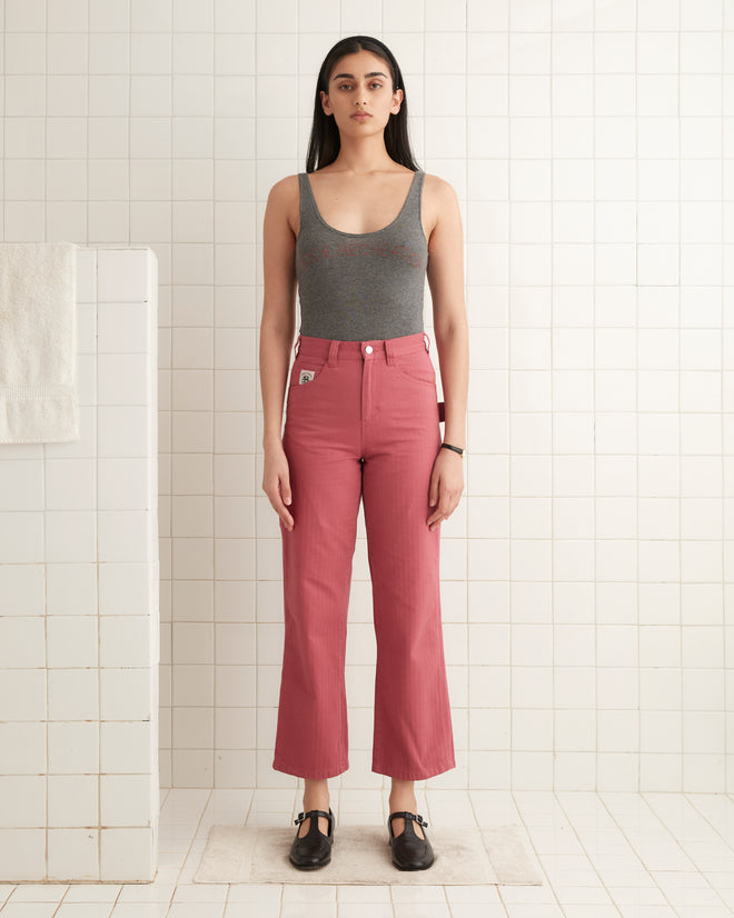 A person in a gray tank top and Knolly Brook Trousers in pink, inspired by 1950s-1960s canvas workwear, stands in a tiled room.