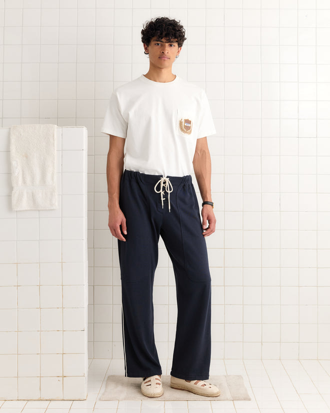 A young man stands in a tiled room with a towel nearby, wearing navy Lace-Front Sweatpants made from heavyweight fabric and a white t-shirt.