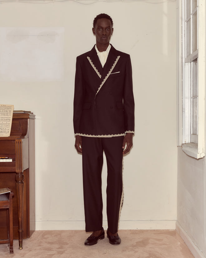 A person in a Lacework Double-Breasted Tuxedo Jacket with gold trim stands indoors by a piano and window, facing forward.