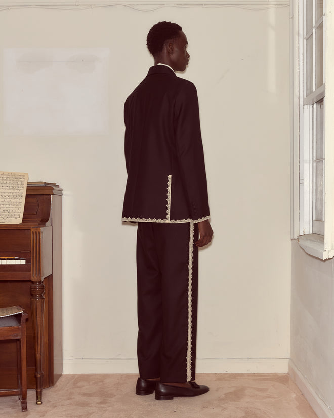 A man in a Lacework Double-Breasted Tuxedo Jacket stands facing a wall near a piano and window.