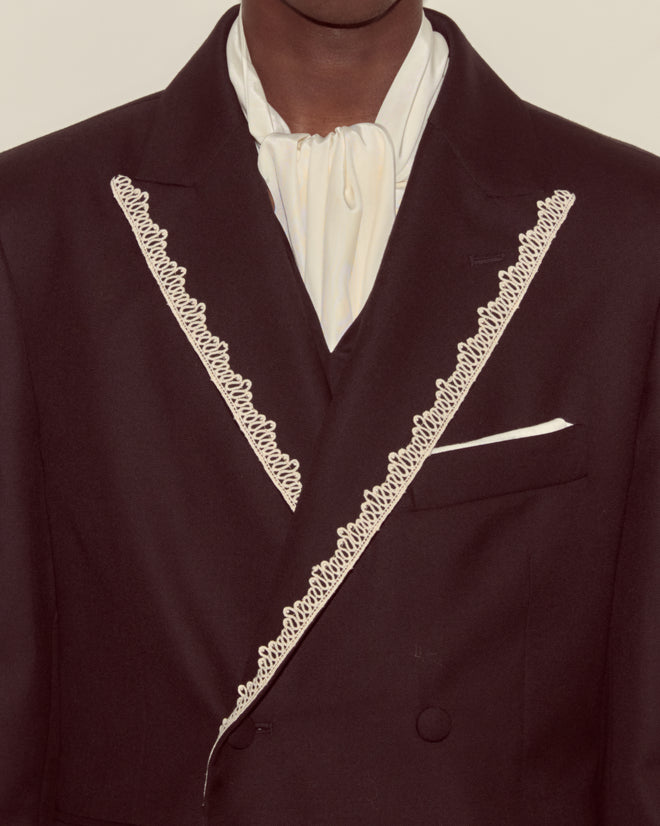A person wearing the Lacework Double-Breasted Tuxedo Jacket with lace trim and a white cravat, shown from shoulders to chest.