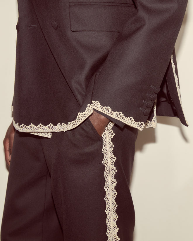 A close-up of the Lacework Double-Breasted Tuxedo Jacket, a dark wool suit featuring cream lace trim along the jacket’s hem, pockets, sleeves, and pants.