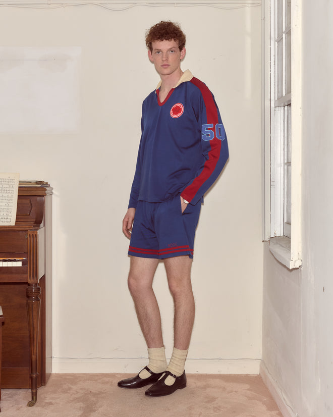 A person in a blue Lakewood Polo and dress shoes stands indoors next to a piano and a window.