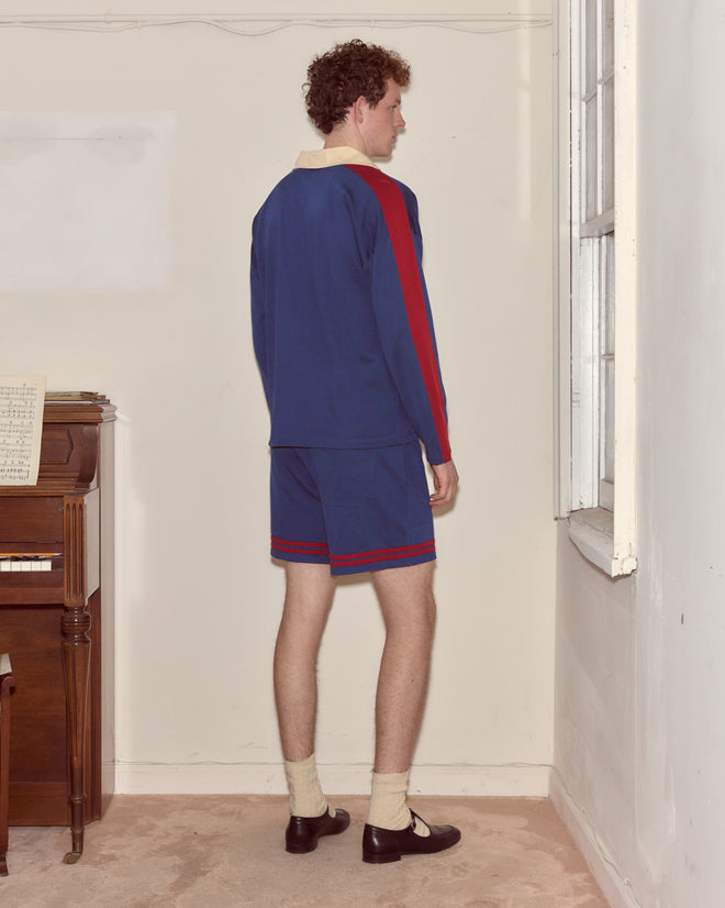A person in Lakewood Polo shorts and a top with red stripes stands facing a corner near a window and piano.