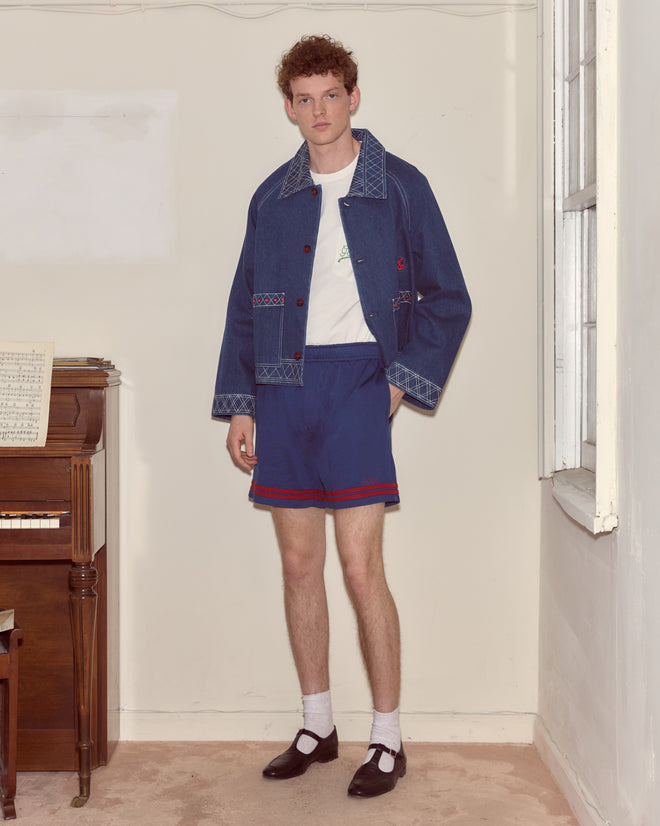 Young man in a blue jacket, Lakewood Shorts, white socks, and black shoes stands indoors next to a piano.