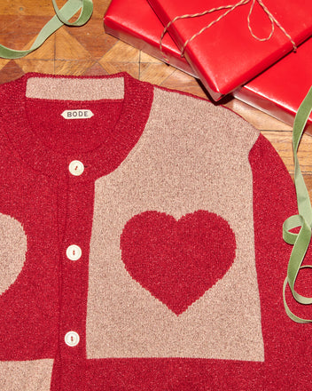 Lamé Hearts Cardigan featuring a red and beige heart pattern in lurex yarn, displayed beside red gift boxes and green ribbon on a wooden surface.