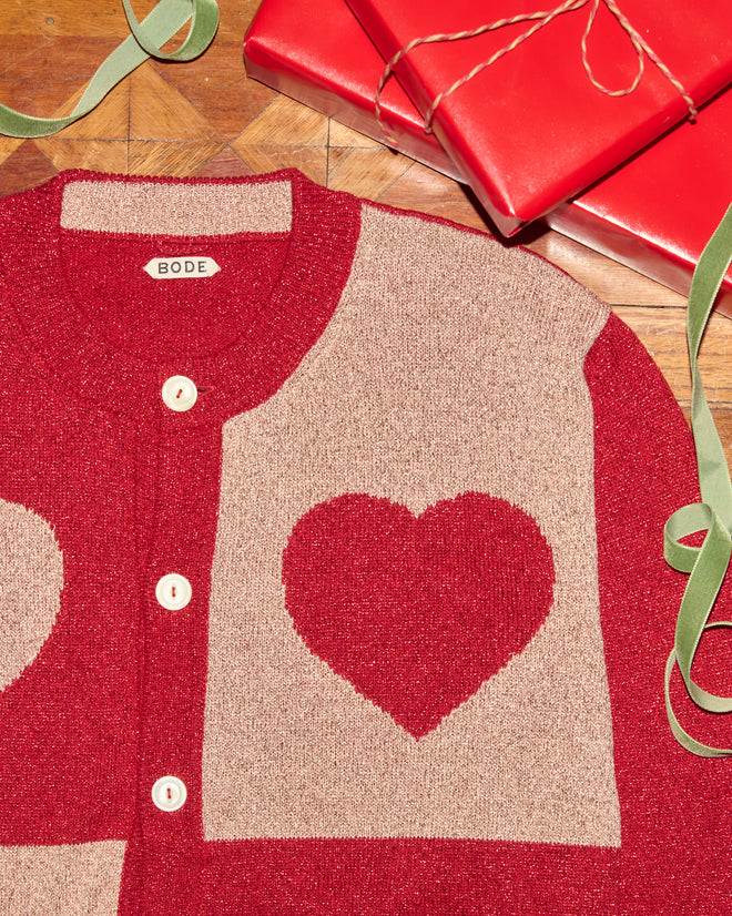 Lamé Hearts Cardigan featuring a red and beige heart pattern in lurex yarn, displayed beside red gift boxes and green ribbon on a wooden surface.